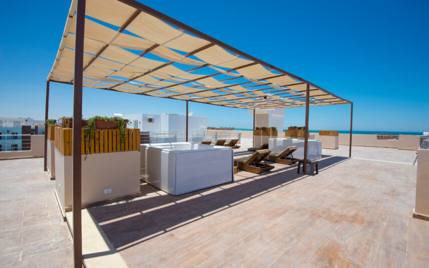 Moderne Dachterrassen Idee mit mediterraner Optik – Auf der Liege mit Sonnenschutz das Wetter genießen – Beispiel mit Whirlpool & bepflanzten Blumenkästen
