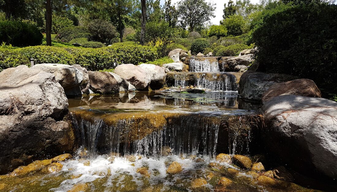 Idee für einen großen Garten - Beispiel mit langen Wasserfall mit großen Steinen umrahmt von immergrünen Hecken