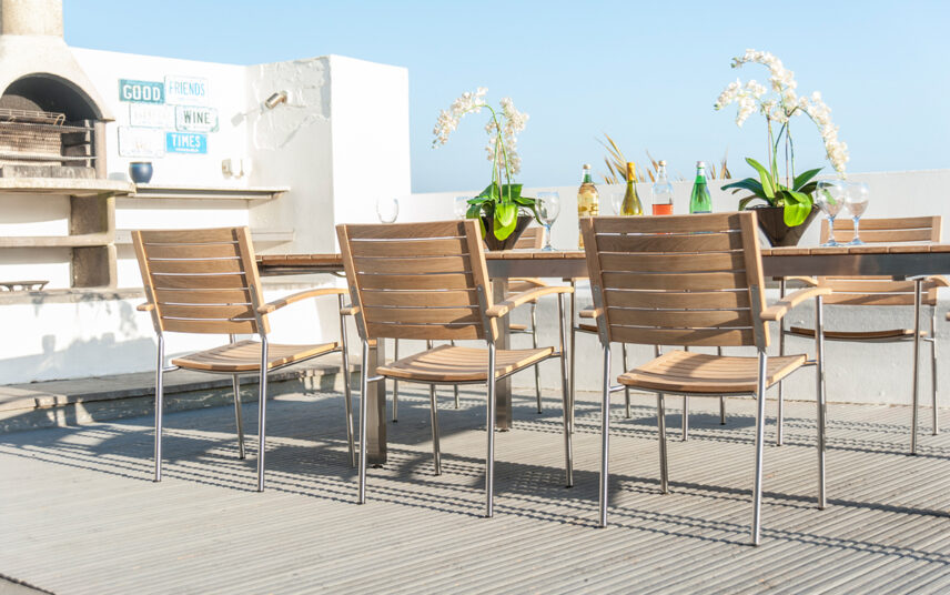 Mediterrane Dachterrasse mit hellen Stühlen und offenem Grillofen – Holzstühle & Holztisch mit Metallakzente – Terrassenpflanzen in Pflanzgefäßen