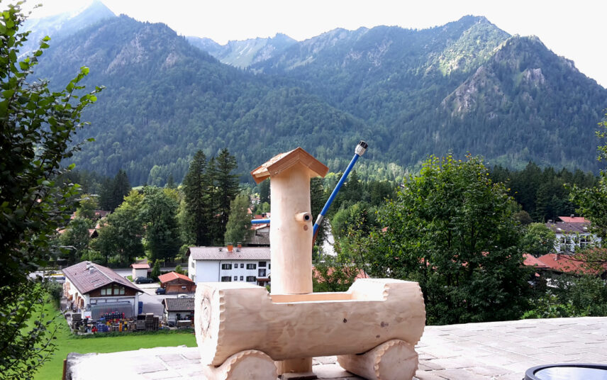 Alpiner Baumstammbrunnen auf einer gepflasterten Terrasse im Garten Idee – ländliche Umgebung mit Bergen – große Wiese im Garten