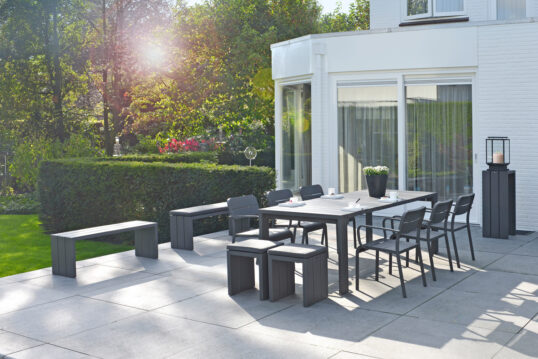 Moderne Terrassengestaltung mit Sitzgruppe – Beispiel mit schwarzen Tisch & schwarzen Stapestühlen – schwarze Bänke & passend dazu ein Beistelltisch mit moderner Gartenlaterne – Hecken & Sträucher