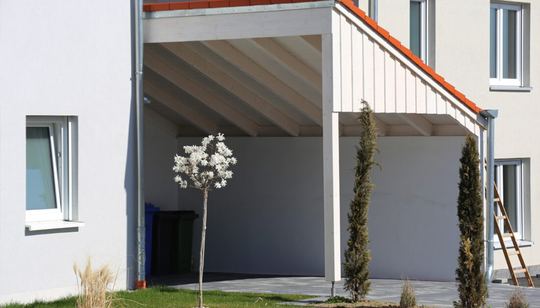 Moderner Anlehn-Carport in weiß mit roten Dachziegeln an der Hauswand als Gartenidee - Vorgarten mit Wiese und kleinen Bäumen
