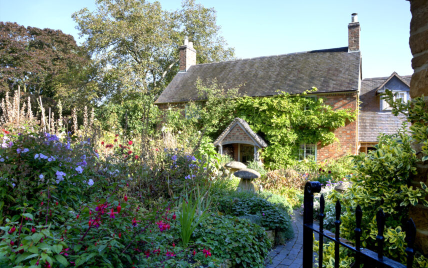 Cottagegarten Idee mit Gartenweg & Gartendekoration – Beispiel mit Dekopilzen aus Stein  bewachsener Hausfassade & Sommerblumen