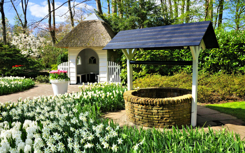 Gartenidee mit märchenhaften Steinbrunnen & weißen Holzgartenhaus – Landhausgarten Beispiel mit Pflanzgefäß als Deko & Gartenpflanzen