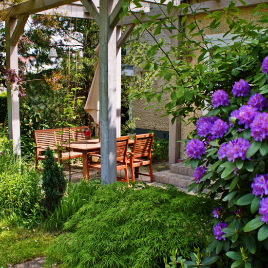 Idee für eine kleine Außenterrassse mit Sitzgruppe aus Holz - Blühender Rhododendron (lila) im Vordergrund - Pergola aus Holz als Gestaltungslement