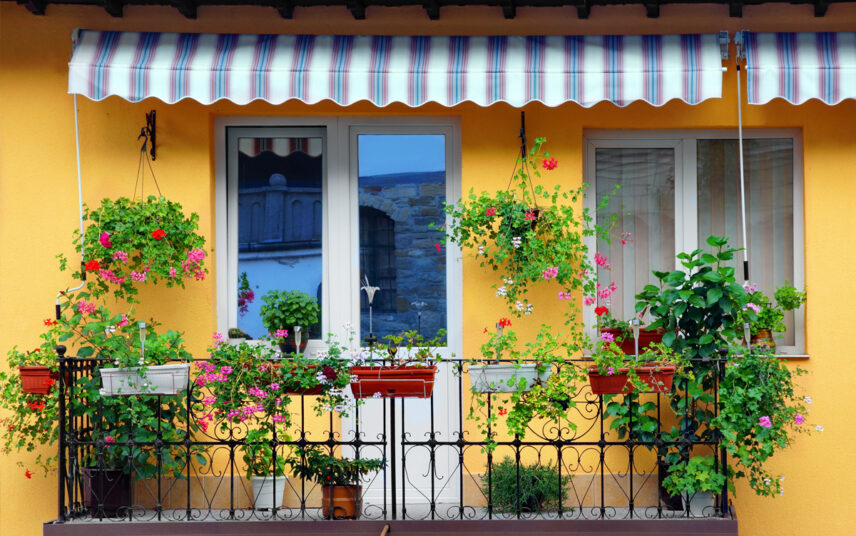 Balkon Idee – Kleiner Balkon an einer orangenen Hauswand dekoriert mit vielen Blumenampeln  Blumenkästen am Balkongeländer & Pflanzgefäße – Balkon Markise als Sonnenschutz