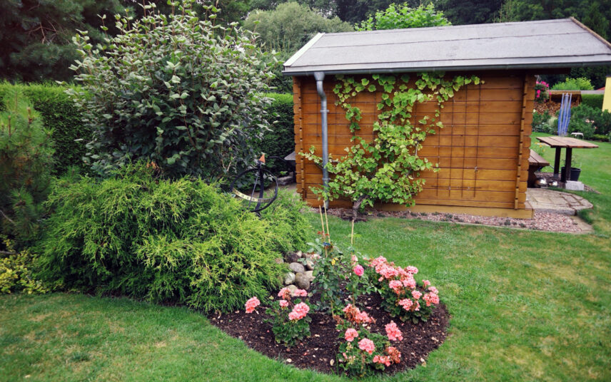 Gestaltungsidee für den Garten mit Rankgittern am Holzgartenhaus – Bepflanztes Beet mit Rosen & Sonnenuhr aus Metall – kleiner Sitzbereich vor dem Gartenhaus