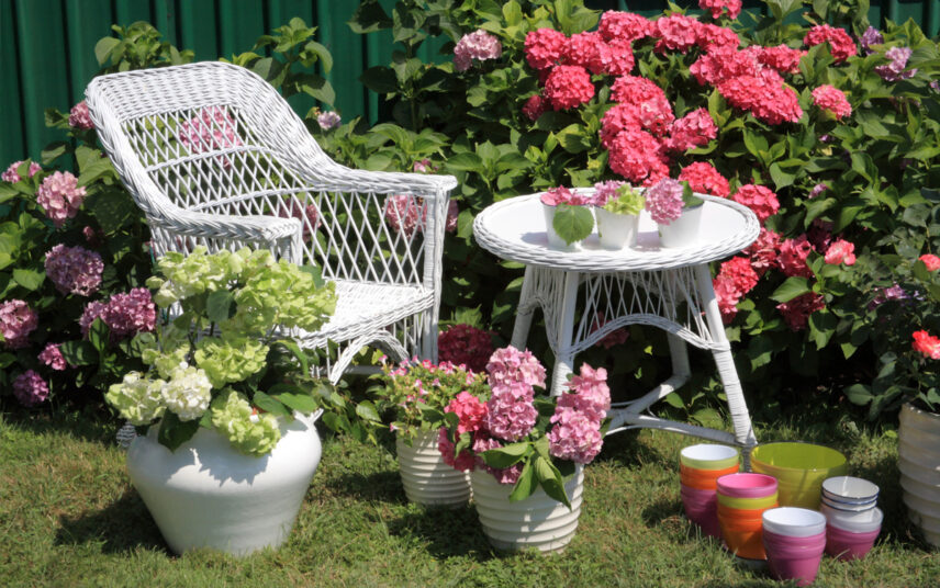 Gestaltungsidee für den Landhausgarten – Weiße Rattansitzgruppe mit Stuhl & Tisch – Beet & Pflanzgefäße mit blühenden Hortensien in rosa & weiß