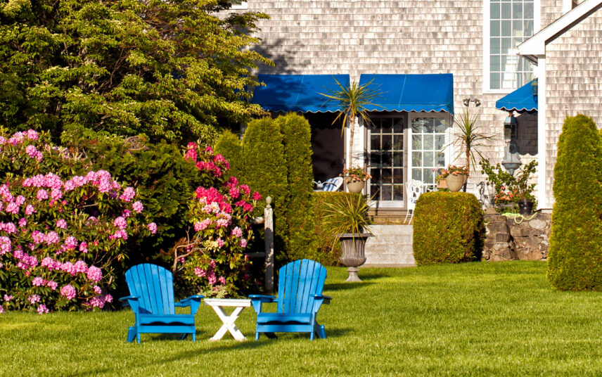 Idee für einen großen Garten – Terrasse & Wiese vor dem Haus  – Bequeme Holzstühle mit Holztisch vor Rhododendron – Pflanzen in Pflanzgefäßen – Blaue Markisen an der Terrasse