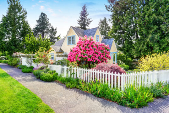 Garten Gestaltungsidee – Kleines gelbes Haus mit weißem Zaun & blühenden Rhododendron – viele Gartenpflanzen am Gartenzaun