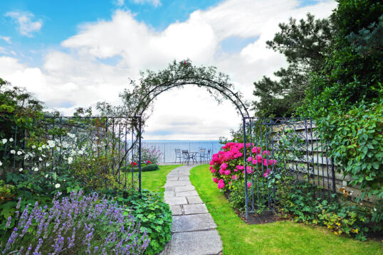 Landhausgarten Idee – Gartenweg mit bepflanzten Rosenbogen aus Metall – Beete mit Lavendel & rosa Hortensien – Sitzgruppe aus Metall am Ende des Gartenwegs
