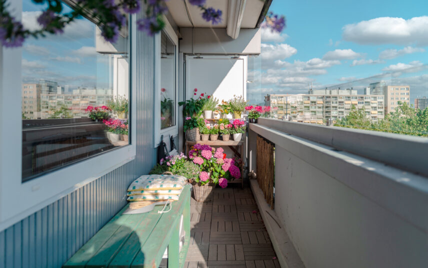 Idee für einen schmalen Balkon mit bemalter Holzbank & Pflanzregal – Blumentöpfe mit rosa Hortensien & weiteren blühenden Blumen – Kissen auf der Bank