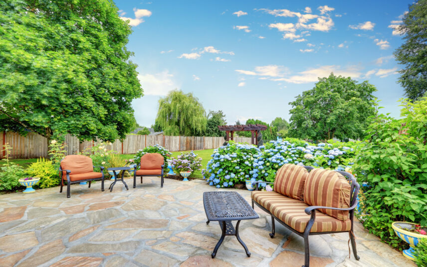Terrassen Idee – Große runde Terrasse mit Bank  Sesseln & Tischen aus Metall – Große blühende Hortensien & Pflanzgefäße mit Blumen – Holzpergola im Hintergrund