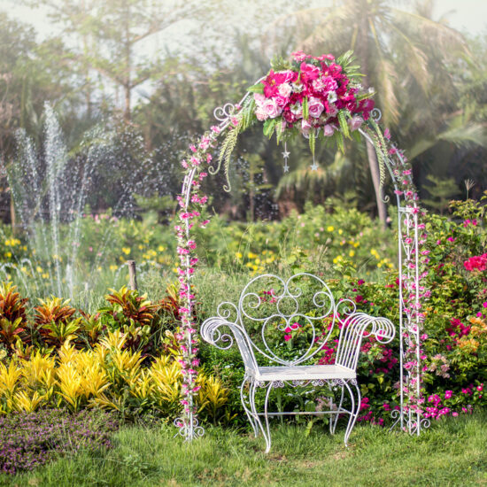 Romantische Gartenidee - Weißer Metallstuhl unter einem dekorierten Rosenbogen mit Strauß - Blumengarten mit rosa Rhododendren - Fontäne im Teich