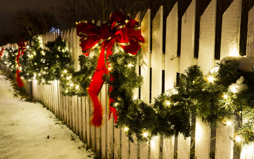 Gartenidee mit Weihnachtsdeko – Beispiel mit weihnachtlicher Dekoration am weißen Gartenzaun – Große rote Schleifen an der Weihnachtsgirlande & Lichterketten