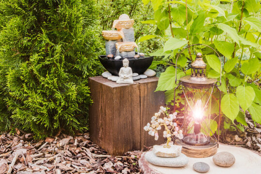 Gartendekoration im asiatischen Stil – Buddha-Skulptur & kleiner Brunnen auf einen Holzhocker – Laterne & Steindekoration