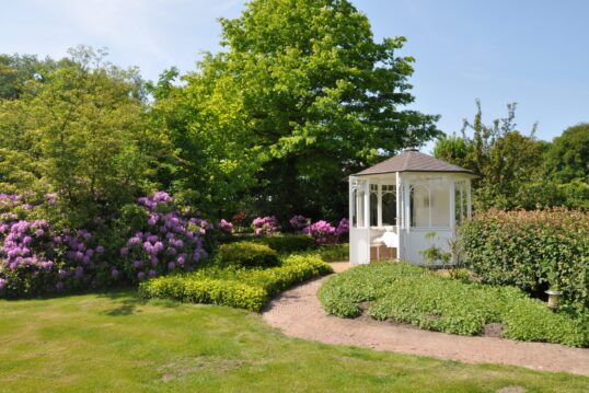 Romantische Gartenidee mit weißen Pavillon – Angelegter Gartenweg mit Pflanzen rechts & links vom Weg – lila & rosa Rhododendren