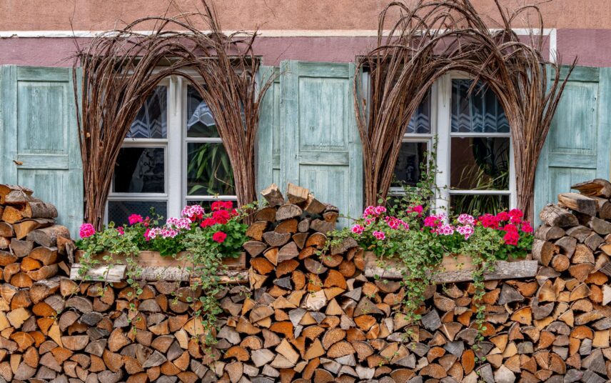 Gestaltungsidee im Landhausgarten – dekorierter Brennholzstabel vor der Hausfassade mit Sommerblumen und getrockneten Weinranken