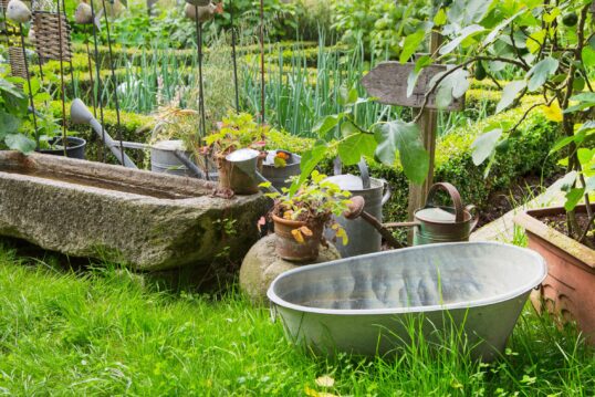 Dekorationsidee Bauerngarten - gefüllter Wassertrog und Zinkgefäße mit Gießwasser für Gemüsebeete & rustikale Gartenstecker aus Stein und Rattan