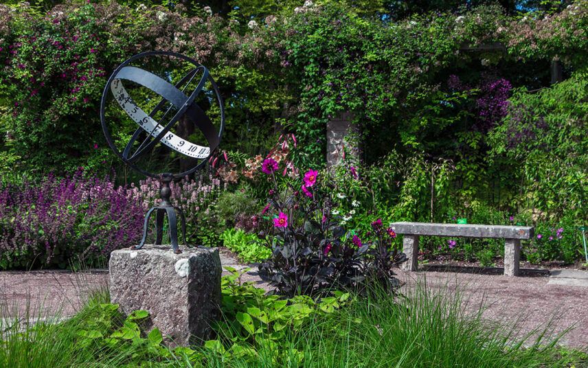 Große Sonnenuhr im Garten auf einem Steinstockel – Steinbank auf dem Kies-Gartenweg