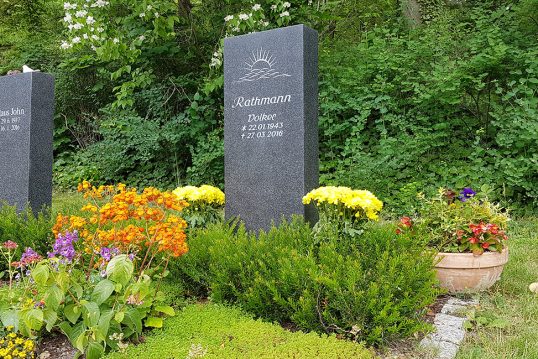 Moderne Grabgestaltung eines Doppelgrabes mit dunkler Granitstele – natürliche Grabeinfassung mit Eibe & Bepflanzung mit einer Mischung aus Bodendeckern und Sommerblumen
