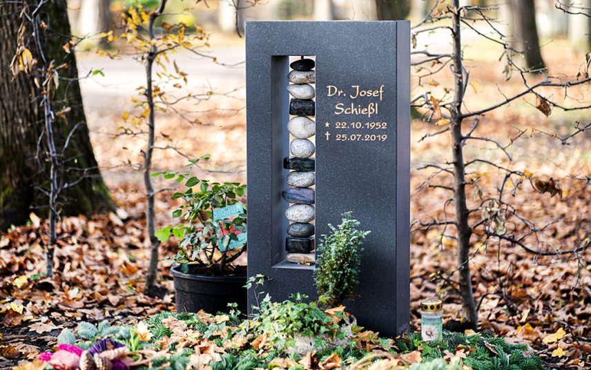 Modern gestalteter Urnengrabstein aus poliertem Granit mit Kieselsteinsäule in einer Öffnung – Wintereindeckung des Grabes mit Tannenreisig