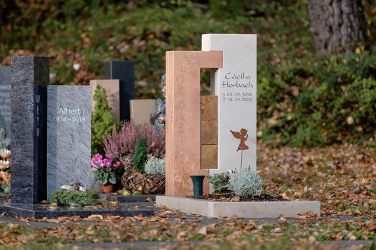 Moderner Urnengrabstein aus hellem Kalkstein und Rotem Travertin mit Holzwürfeln verbunden – winterliche Grabgestaltung mit Stauden und Gesteck