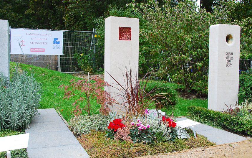 Moderner Grabstein mit Einfassung aus rotem Granit auf einem Urnengrab mit Loch und Bronzeplastik – Sommerliche Grabbepflanzung und Grabschmuck