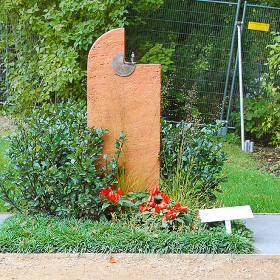Modern gestalteter Grabstein aus rotem Travertin- halbrund mit kleinem Bronzerelief - Grabbepflanzung mit Stechpalme & Zier-Paprika