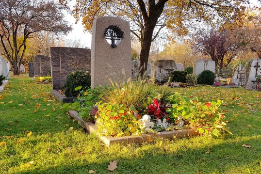 Moderner Grabstein mit Einfassung aus rotem Granit auf einem Urnengrab mit Loch und Bronzeplastik – Sommerliche Grabbepflanzung und Grabschmuck