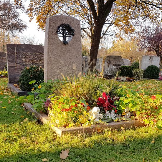 Moderner Grabstein mit Einfassung aus rotem Granit auf einem Urnengrab mit Loch und Bronzeplastik - Sommerliche Grabbepflanzung und Grabschmuck