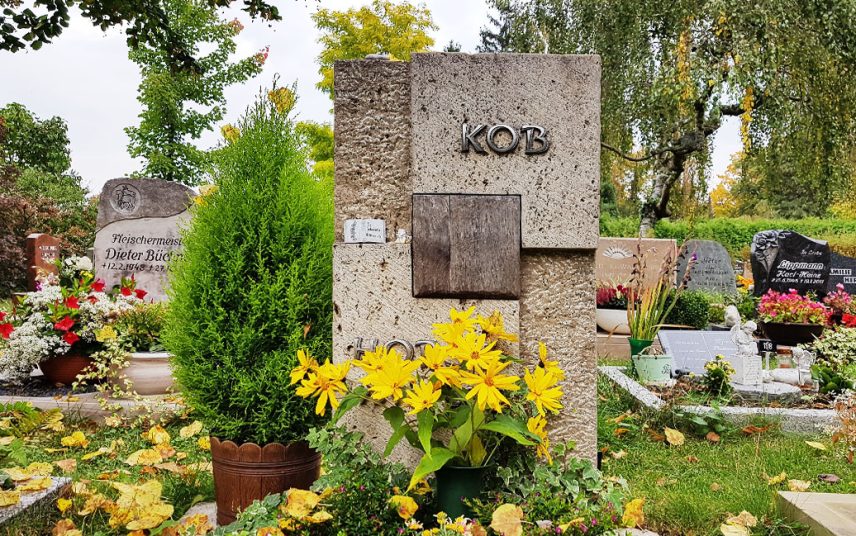 Moderner geometrischer Urnengrabstein aus Travertin und Eichenholz – Grabgestaltung mit Blumen im Sommer