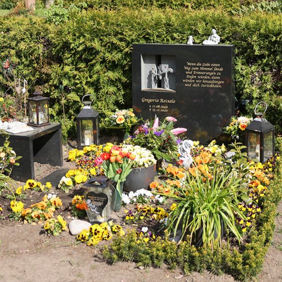 Moderner Doppelgrabstein aus schwarzem poliertem Granit mit Öffnung und zwei Bronzeengeln - Üppige Grabgestaltung mit Frühlingsblumen und Grabdekoration