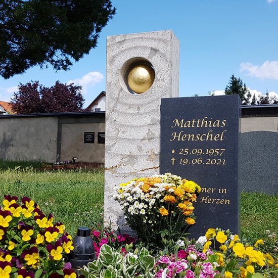 Auffälliges Urnengrab mit modernem Grabstein aus einem Materialmix aus Kalkstein & Granit und einer vergoldeten Kugel - Grabgestaltung mit üppig blühenden Frühlingsblumen