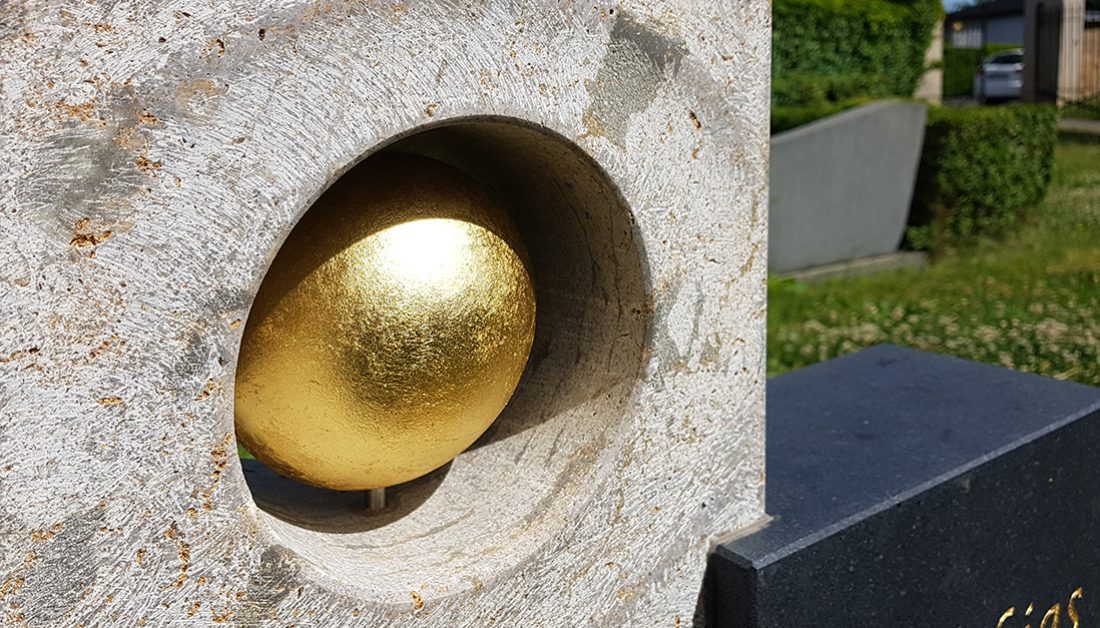 Auffälliges Urnengrab mit modernem Grabstein aus einem Materialmix aus Kalkstein & Granit und einer vergoldeten Kugel – Grabgestaltung mit üppig blühenden Frühlingsblumen
