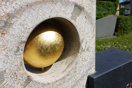 Auffälliges Urnengrab mit modernem Grabstein aus einem Materialmix aus Kalkstein & Granit und einer vergoldeten Kugel – Grabgestaltung mit üppig blühenden Frühlingsblumen