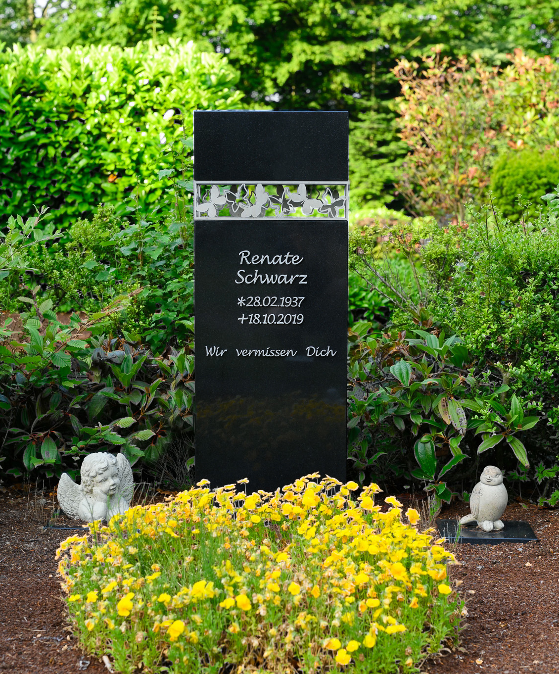 Einfache Grabgestaltung mit schwarzem Grabstein und weißen Bodendeckern. Rechts und links vom Grabstein stehen kleine Grabfiguren.