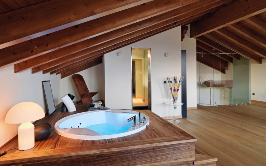Wohnidee – Relaxbereich zu Hause unter dem Dach mit Whirlpool – Glasbeistelltisch mit Dekoration – Holzliege & Tischlampe – runde Wandleuchten an der weißen Wand