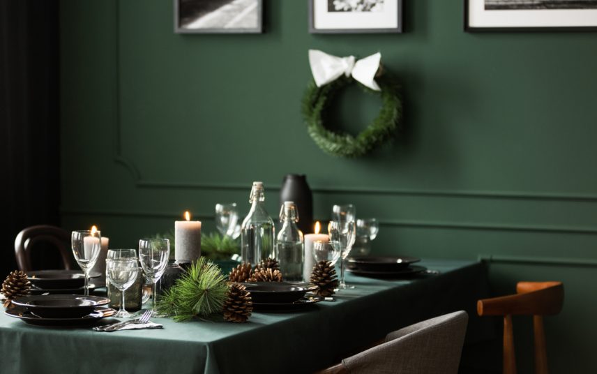 Idee für die Tischdeko zu Weihnachten – Weihnachtlich geschmückter Esstisch vor einer grünen Wand – Grüne Tischdecke & graue Kerzen – Tischdeko mit Tannenzapfen & Zweigen