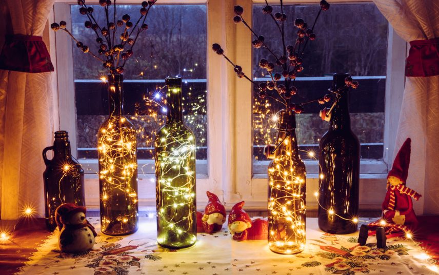 Idee mit Weihnachtsdeko auf der Fensterbank Innen – Glasflaschen mit Lichterketten & Weihhnachtsfiguren auf einer Tischdecke